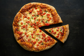 freshly cooked simple homemade pizza with cheese, tomatoes and green onions on thick dough cut into slices lies on black wooden background. top view with copy space