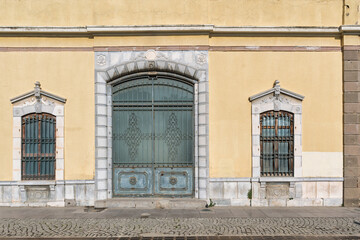 Exterior view of an ornate building facade featuring a large arched gate, two arched windows, yellow wall, and stone details above a cobblestone sidewalk