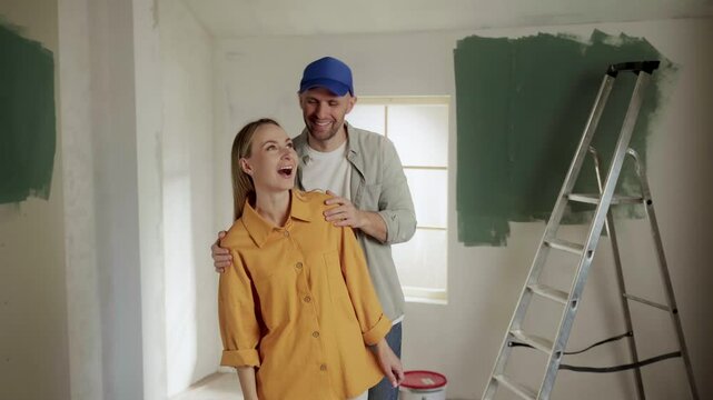 Painter is covering a woman's eyes, creating a surprise during their home renovation project, with a partially painted green wall and a ladder in the background