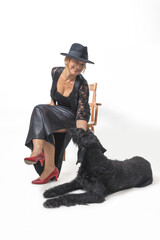 Middle aged woman is sitting on a chair in a studio. A Big Black Schnauzer dog is lying at her feet.