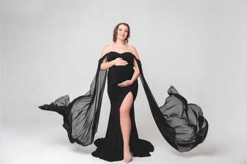 Beautiful pregnant young girl in a black long dress on a white background in a photo studio....