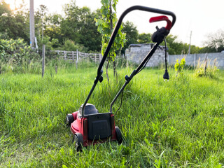 Red electric lawn mower cutting green grass in garden at sunset