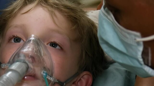 Young blond boy with an oxygen mask lies on a surgical table in an operating room surrounded by medical staff during a procedure.