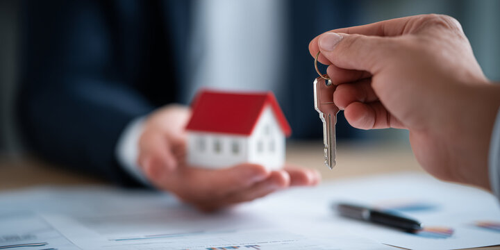 Hands exchanging a house key and miniature home model, business context, signing documents in background, real estate deal, mortgage agreement concept