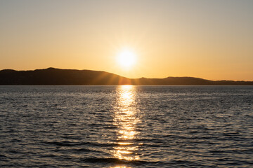 A golden sunset over the calm waters of Norway, reflecting on the surface and illuminating the silhouettes of the distant coast.