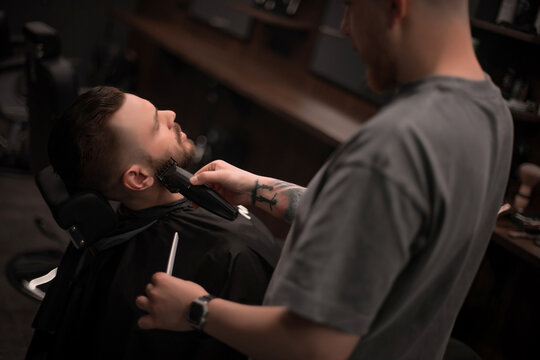Barber trims client's beard with electric clippers in modern barbershop. Close-up of grooming session with professional barber and relaxed male customer in stylish interior..