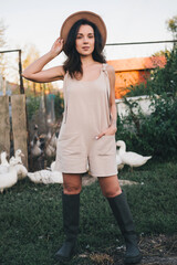A beautiful woman in a light jumpsuit with a hat stands near a paddock with geese village. tinting