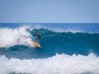Male surfer in a wetsuit riding a smooth, clean blue wave with an orange board. The wave curls...