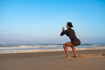 Athletic woman in black sportswear doing squat exercise on sandy beach at sunrise, facing the...