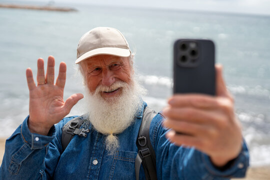 cheerful elderly man with a white beard, wearing a beige cap and denim shirt, waves while taking a selfie with his smartphone on a sunny beach with ocean waves in the background