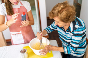 Grandmother whisking ingredients while granddaughter is holding essence bottle in kitchen