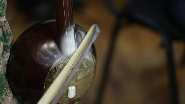A close-up shot captures a musician's hands skillfully playing a traditional kamancheh, a spike fiddle, drawing the bow across its strings in a dynamic and focused musical performance.