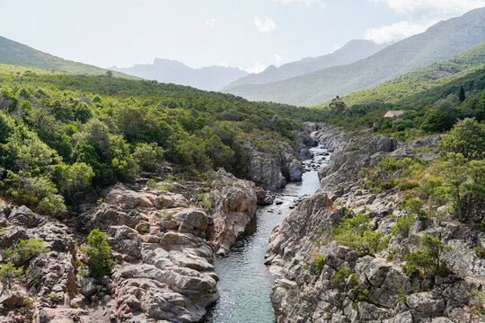A scenic river flows through a rocky, green landscape, with mountains in the distance. Vall&eacute;e du Fango - U Valle di u Fangu, Corsica.