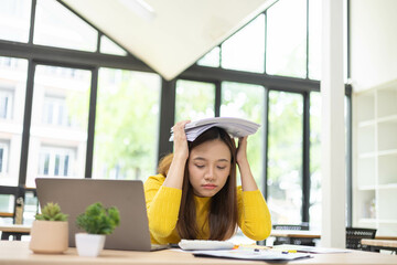 A woman is sitting at a desk with a laptop and a stack of papers. She is looking at the laptop and...