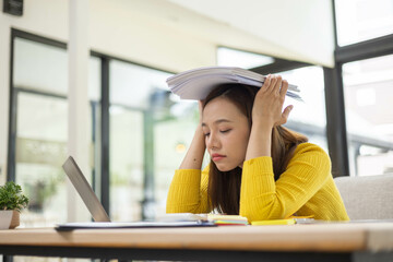 A woman is sitting at a desk with a laptop and a stack of papers. She is looking at the laptop and...