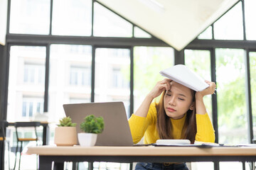 A woman is sitting at a desk with a laptop and a stack of papers. She is looking at the laptop and...