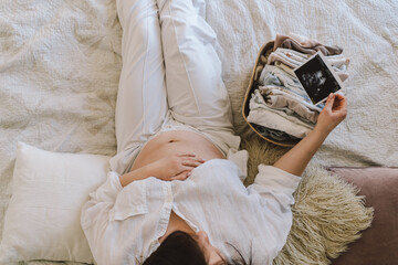 A pregnant woman lies on a bed with an ultrasound image and a basket of baby clothes. The moment of waiting and bonding at home.