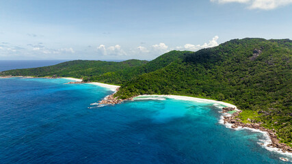 A beach cove with white sand surrounded by lush green hills and clear blue ocean water. Seychelles, La Digue.