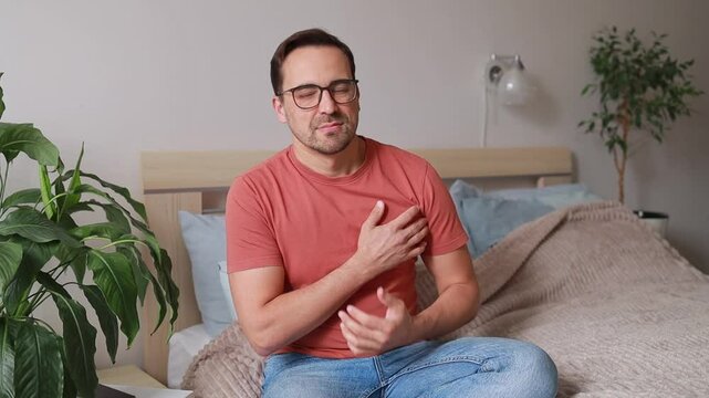 A man sits on a bed, suddenly clutching his chest in pain. His face expresses fear and anxiety. Heart attack, infarction. Concept: cardiology, health.