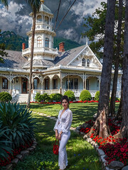 woman in white posing in front of Victorian style wooden mansion with lush lawn on a bright partly cloudy day