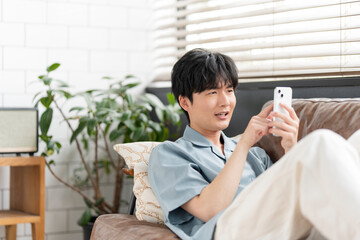 Obraz premium ソファーに座りリラックスしながらスマートフォン の画面を見ている若い日本人男性。