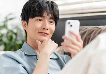 ソファーに座りリラックスしながらスマートフォン
の画面を見ている若い日本人男性。
