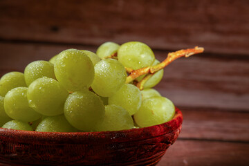 Cluster of ripe green grapes with drops of water, served in a carved wooden bowl on a wooden table.