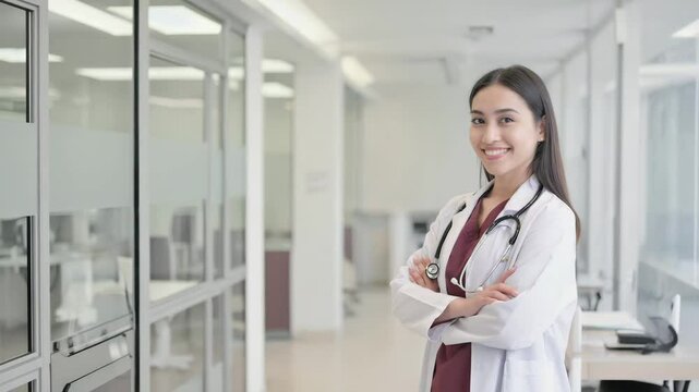 A confident and smiling woman medical professional stands in a bright hospital corridor with crossed arms