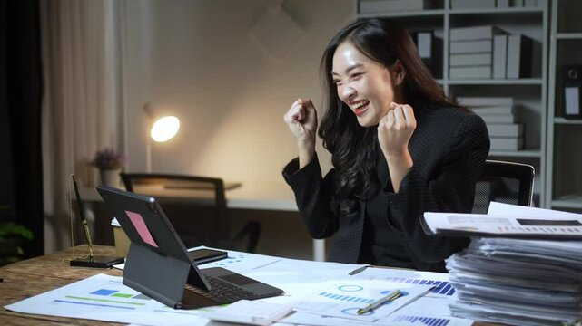 A woman is sitting at a desk with a laptop and a stack of papers. She is typing on the laptop and smiling