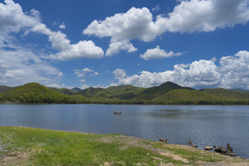 Serene lake surrounded by lush green hills under bright blue sky with fluffy white clouds, creating peaceful atmosphere