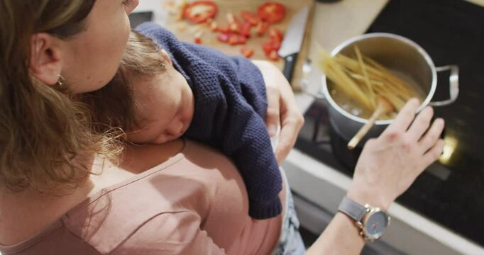 Video of caucasian mother with newborn baby in baby carrier cooking in kitchen