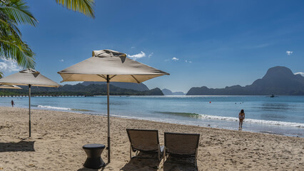 A sunny day, a tropical beach. Two sun loungers under sun umbrellas. The waves of the calm turquoise ocean are foaming. Silhouettes of people. Mountains on the horizon.Palm leaves against the blue sky