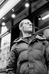 Young man wearing sunglasses and puffer jacket looking up in monochrome analog film style