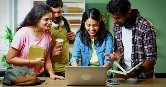 Indian college students using laptop for completing group assignment, standing with books and notes, working together in friendly college library environment, focused on academic teamwork