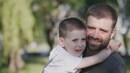 Fototapeta premium A father smiles while his young son hugs him tightly in a sunny park setting, showcasing their close bond and joyful experience together in the pleasant weather.
