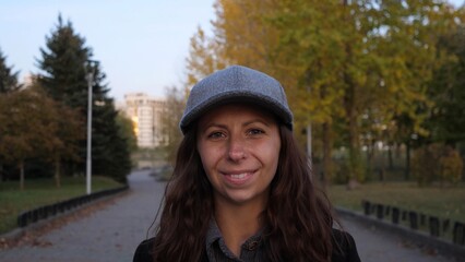 A woman with long, curly hair wearing a gray cap enjoys a relaxed moment while walking on a serene path surrounded by vibrant autumn leaves and peaceful trees.