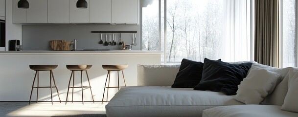 Sunlit minimalist living space featuring a white sectional sofa with dark cushions, three wooden bar stools at a kitchen island, and large windows showcasing a wintery landscape