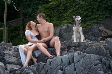 A young girl and a guy are sitting on the stone steps of a rock overgrown with vines, and they smile at each other, and a faithful dog is sitting next to them.