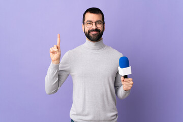 Adult reporter man with beard holding a microphone over isolated purple background showing and lifting a finger in sign of the best