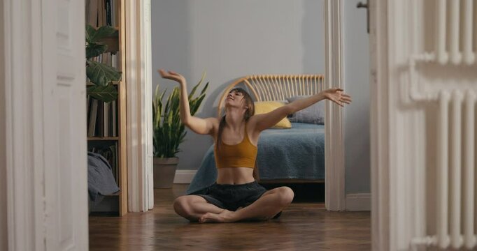 Happy girl in yellow top sitting on floor and dancing with hands during fun contemporary dance improvisation at home in modern apartment