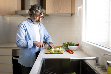 Mature man cutting lettuce and vegetables in kitchen to prepare fresh salad for healthy lunch. Copy...