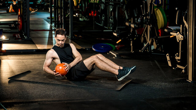 Focused young man performing Russian twist with medicine ball on yoga mat at gym, strengthening core and improving balance during functional workout session.