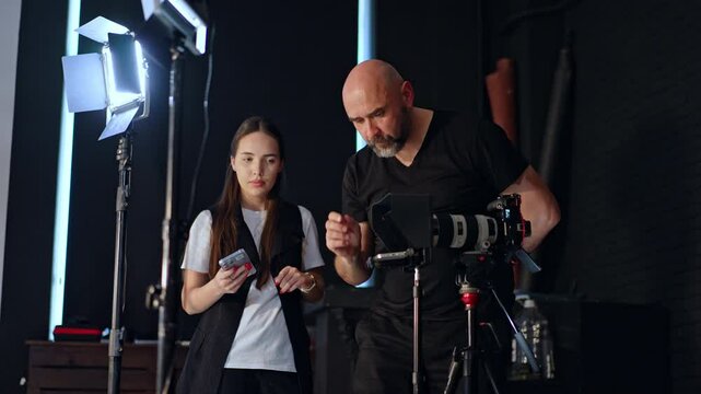 Bearded bald photographer stands at the camera on tripod. Cameraman sets the video prompter. Long-haired girl stands nearby focused on her phone.
