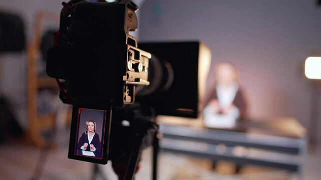 Professional camera recording a female reporter sitting at desk at blurred backdrop. Close up. Blogging, tv program, newsreader concept.