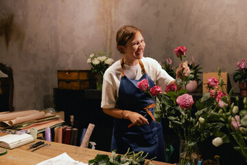 Middle-aged female florist in apron arranging fresh flowers for bouquet in the flower shop. Small...