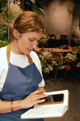 Middle-aged female florist in apron arranging calling on the smart phone. She is arranging...