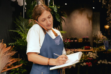 Middle-aged female florist in apron arranging calling on the smart phone. She is arranging...