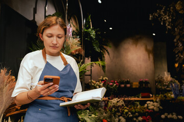 Middle-aged female florist in apron arranging calling on the smart phone. She is arranging...