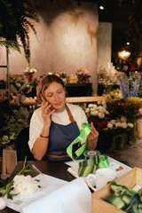 Middle-aged female florist in apron arranging calling on the smart phone. She is arranging...
