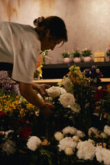 Middle-aged female florist in apron arranging fresh flowers for bouquet in the flower shop. Small...